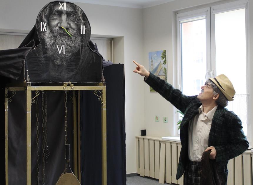 1.Aktor w kapeluszu wskazujący palcem na zegar z nadrukiem starca D