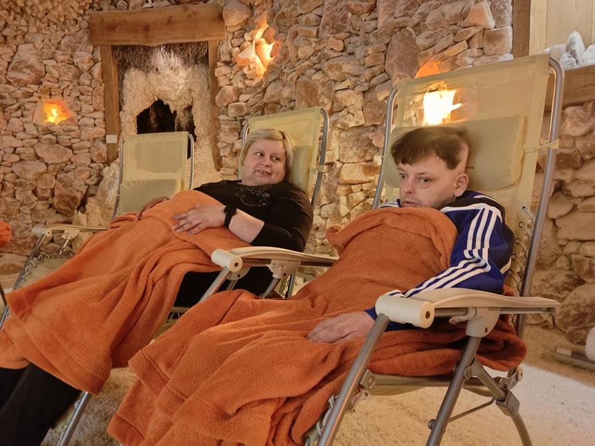 Opiekunka oraz uczestnik siedzący w leżakach pod kocami na tle solnej ściany 
