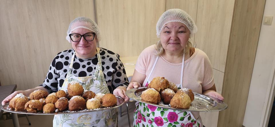 Dwie uczestniczki w fartuchach kuchennych trzymające upieczone pączki na srebrnych tacach 