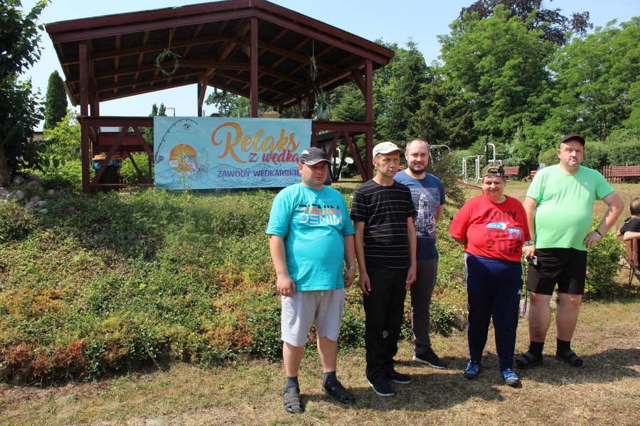 Uczestnicy pozują do zdjęcia grupowego na tle drewnianej wiaty z banerem.