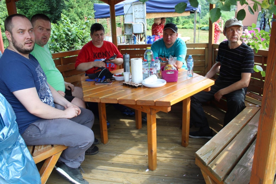 Uczestnicy wraz z terapeutą siedzą na ławce wewnątrz drewnianej wiaty. 