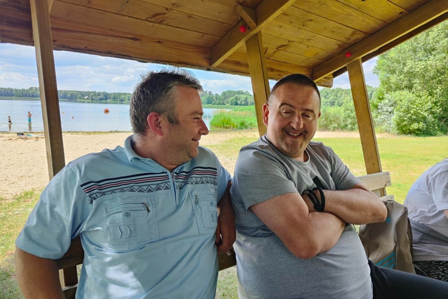 2.Uczestnik oraz kierowca w drewnianej altanie uśmiechający się na tle jeziora