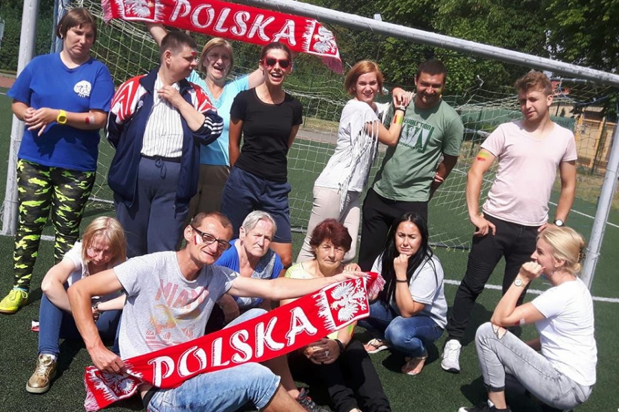 Grupa uczestników wraz z terapeutami trzymająca szaliki Polski na tle bramki 