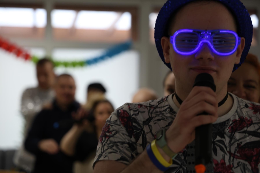 2.Uczestnik z mikrofonem w kolorowych okularach i kapeluszu na tle tańczących osób