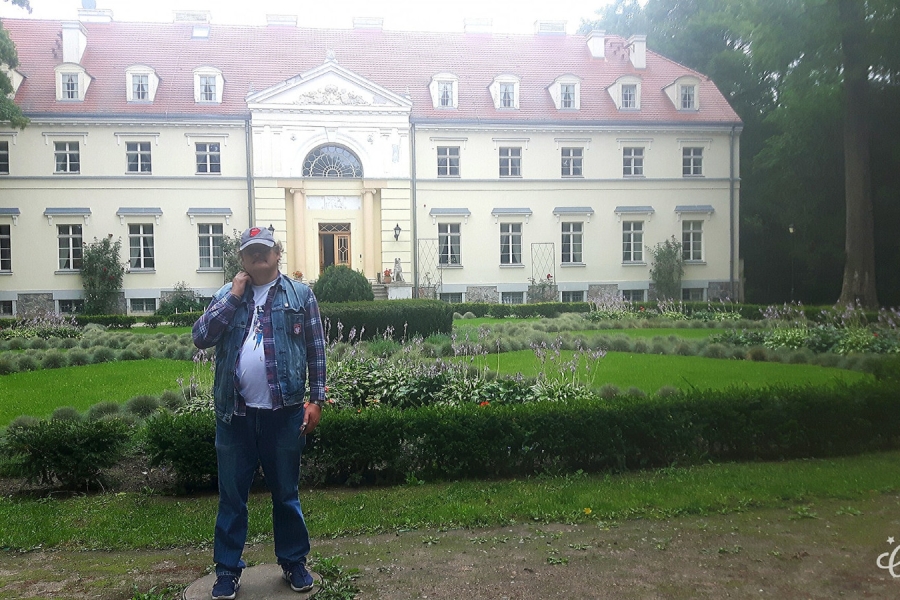 Uczestnik stoi wśród zielni na tle pałacu.