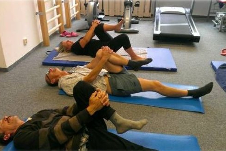 Uczestnicy leżący na karimatach trzymają złączonymi rękoma zgięte kolana. 