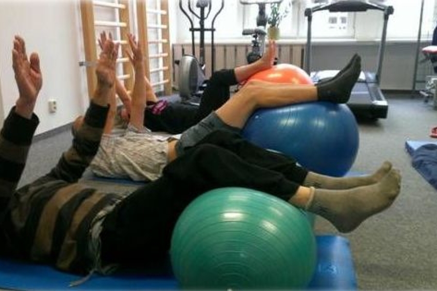 Uczestnicy leżą na karimatach trzymając wyprostowane ręce w górze i zgięte nogi na piłkach 