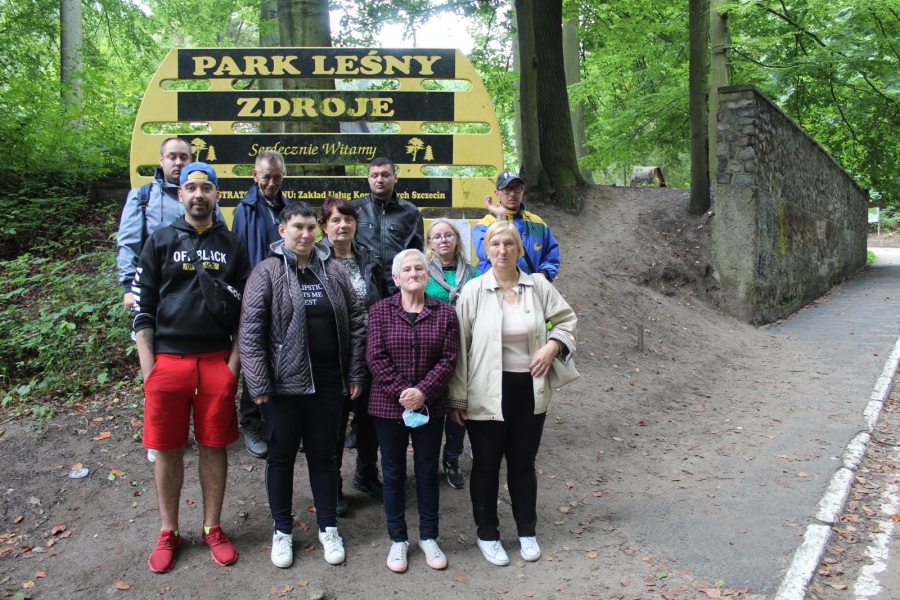 1 – Grupa Uczestników z terapeutą na tle tablicy z napisem Park Leśny Zdroje.