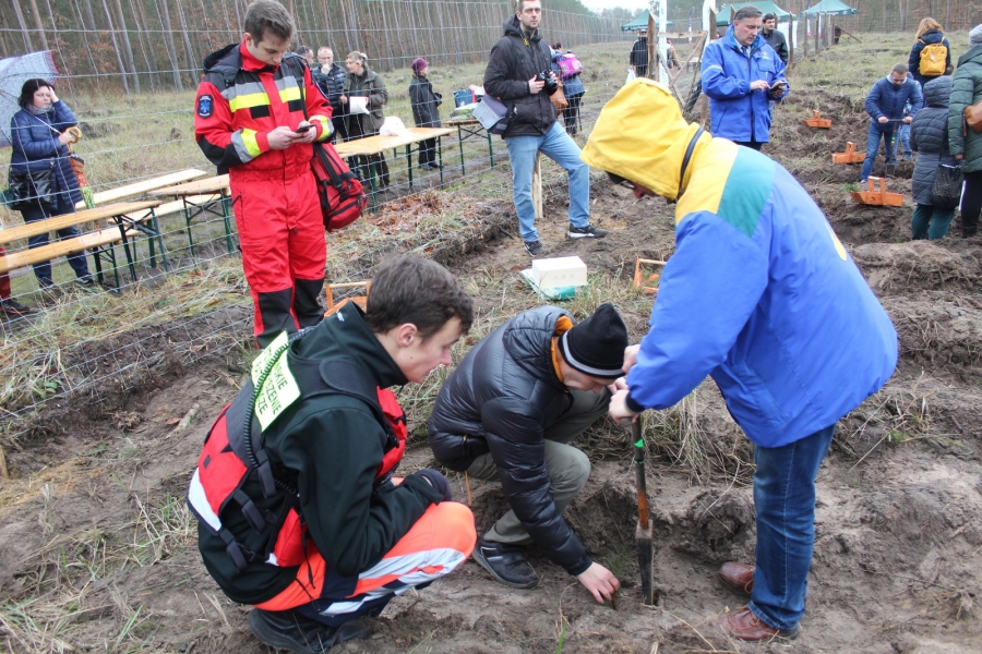6 – Uczestnicy wraz z ratownikiem medycznym sadzą drzewka na tle zaproszonych gości.